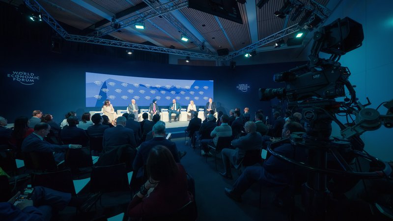 A television camera overlooks a packed audience and expert panel on a blue-branded stage during the World Economic Forum in Davos, Switzerland.